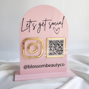 May include: A light pink arched acrylic sign featuring the black text Let's get social with a heart icon. It includes a gold Instagram logo and a square QR code with a gold border, plus the black text @blossombeautyco on a rectangular base.