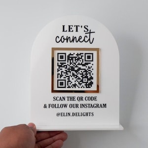 May include: A white arched sign featuring the black text LET'S connect at the top, a central square QR code framed in gold, and the black text SCAN THE QR CODE & FOLLOW OUR INSTAGRAM followed by the handle @ELIN.DELIGHTS at the bottom.