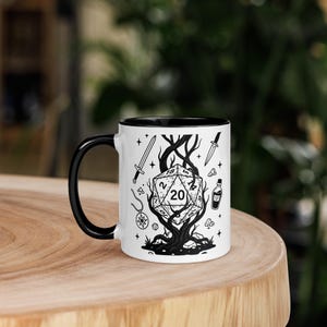 May include: White ceramic mug with a black interior and handle. The mug features a black and white illustration of a D20 die, swords, a potion bottle, and other fantasy-themed elements. The design is centered on the mug.
