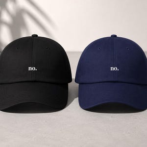 May include: Two baseball caps, one black and one navy blue, each featuring the word no. embroidered in white lowercase letters on the front center. Both caps have a structured crown, a curved brim, and a button at the top of the dome.