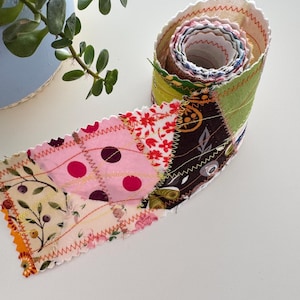 Può includere: Una striscia di tessuto arrotolata con un design patchwork. La striscia presenta vari tessuti fantasia in rosa, verde, marrone e stampe floreali. I bordi sono smerlati e cuciti con filo a contrasto, creando un aspetto artigianale.