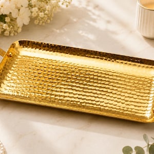 Könnte beinhalten: Ein rechteckiges, goldfarbenes Serviertablett mit gehammerter Textur und integrierten Griffen. Das Tablett ist leer und liegt auf einer hellen Oberfläche, mit einer brennenden Kerze und Blumen im Hintergrund. Dekorative Wohnkultur.