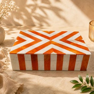 May include: A rectangular storage box featuring a geometric pattern of alternating white and vibrant orange stripes. The lid displays a chevron-like design meeting at the centre, while the sides are decorated with vertical stripes in the same colours.