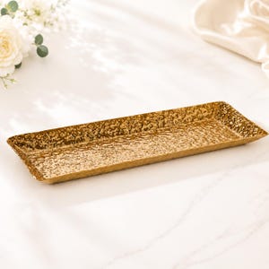 May include: A rectangular gold tray with a textured surface. The tray has slightly raised edges and a hammered metal appearance. The tray is empty and sits on a white surface.