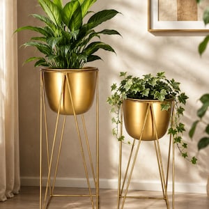 Op de afbeelding: Twee goudkleurige metalen plantenbakken met gladde, ronde kommen op hoge, dunne, geometrische draadstandaards. De hogere plantenbak bevat een groene bladplant, terwijl de kortere plantenbak een hangende groen-witte klimop bevat.