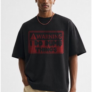 Pode incluir: Uma t-shirt preta de manga curta com um gráfico retangular vermelho de sinal de aviso no peito. O design inclui um ícone de ponto de exclamação e o texto WARNING I DO DUMB THINGS numa fonte preta com efeito de gotejamento.