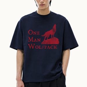May include: A dark navy blue short-sleeved t-shirt featuring a red graphic print of a wolf howling on a rock. The text "ONE MAN WOLFPACK" is printed in red capital letters to the left of the wolf graphic on the front of the garment.