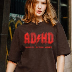 Pode incluir: Uma t-shirt de manga curta castanho-escura com uma impressão gráfica vermelha. O texto diz ADHD com um relâmpago no centro, seguido pelo texto HIGHWAY TO... HEY LOOK A SQUIRREL! numa fonte angular, a negrito e com aspeto desgastado.