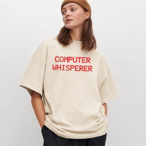 May include: An oversized beige t-shirt featuring the text "COMPUTER WHISPERER" printed in a red, pixelated, blocky font across the chest. The garment has short sleeves, a crew neckline, and a loose, relaxed fit, constructed from a solid-coloured fabric.