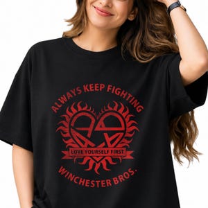 May include: A black short-sleeved t-shirt featuring a red graphic print of a heart with flames. The text reads ALWAYS KEEP FIGHTING, LOVE YOURSELF FIRST, and WINCHESTER BROS. The design has a distressed, weathered texture throughout the red ink.