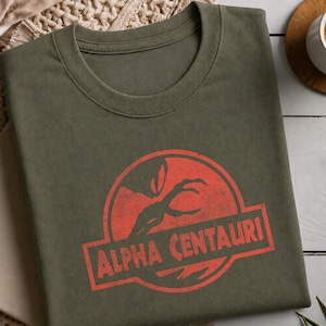 May include: An olive green crew neck t-shirt featuring a distressed red circular graphic. The design depicts a clawed silhouette within the circle, with the text ALPHA CENTAURI printed in a bold, block font across the center of the graphic.
