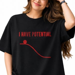 Op de afbeelding: Een zwart t-shirt met korte mouwen en de rode tekst I HAVE POTENTIAL gedrukt boven een rode afbeelding van een bal die op een aflopende lijn rust, wat het concept van potentiële energie illustreert in een eenvoudig en duidelijk ontwerp.