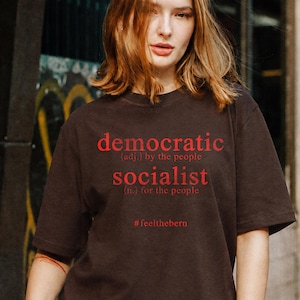 May include: A dark brown short-sleeved t-shirt featuring red text that reads: democratic (adj.) by the people, socialist (n.) for the people, and the hashtag #feelthebern printed in a centred, stacked layout on the front of the garment.