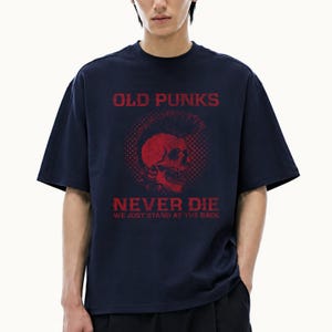 Op de afbeelding: Een marineblauw t-shirt met korte mouwen en een rode afbeelding van een schedel met een hanenkam. De tekst OLD PUNKS NEVER DIE WE JUST STAND AT THE BACK staat in een verweerd rood lettertype boven een cirkelvormig stippenpatroon.
