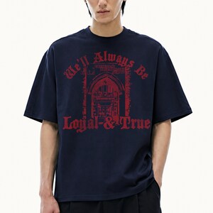 Könnte beinhalten: Ein dunkelblaues T-Shirt mit kurzen Ärmeln und einem roten Grafikdruck auf der Brust. Das Design zeigt einen Torbogen mit dem Text "We'll Always Be" darüber und "Loyal & True" darunter in einer fetten, gotischen Schrift auf der Vorderseite.