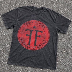 Op de afbeelding: Een antracietgrijs t-shirt met korte mouwen en een cirkelvormige rode afbeelding op de borst. Het ontwerp bevat een zwarte ster, de letters FF en de tekst Fringe Division Department of Defense in een verweerde, versleten stijl.