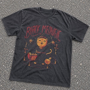 May include: A charcoal gray short-sleeve t-shirt featuring a graphic print of a smiling cat wearing a robe. The text "BLACK MEOWGIC" is printed above the cat, which is surrounded by illustrations of a book, candles, daggers, and a potion bottle.