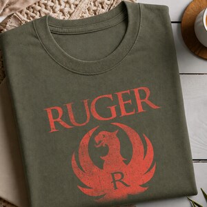 Peut inclure: Un t-shirt à col rond vert olive arborant le mot RUGER imprimé dans une police rouge vieillie au-dessus d'un emblème graphique rouge représentant un aigle stylisé avec la lettre R centrée en bas, le tout avec une texture usée et vintage.