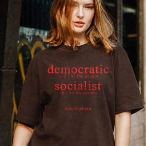 May include: A dark brown short-sleeved t-shirt featuring red text that reads: democratic (adj.) by the people, socialist (n.) for the people, and the hashtag #feelthebern. The shirt has a crew neckline and a standard fit with short sleeves.