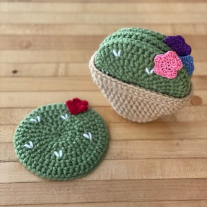 May include: A set of crocheted items including a round green cactus-shaped coaster with a red flower and a green cactus in a tan pot, featuring small white embroidered stitches and pink, purple, and blue crocheted flowers on the cactus top.