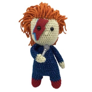 May include: Crocheted doll resembling a rock star, with orange yarn hair and a red and blue lightning bolt on the face. The doll wears a blue jumpsuit and red shoes, holding a silver microphone. A unique handmade item.