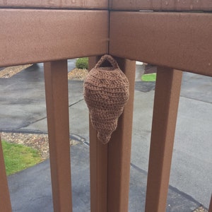 Puede incluir: Una casita de p&aacute;jaros de ganchillo marr&oacute;n colgada de una barandilla de madera.