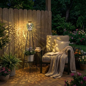 May include: A solar-powered outdoor lantern with a decorative metalwork design, casting intricate light patterns. The lantern is on a black tripod stand, next to a chair with a blanket and a small side table. The scene is set on a wooden deck with potted plants.