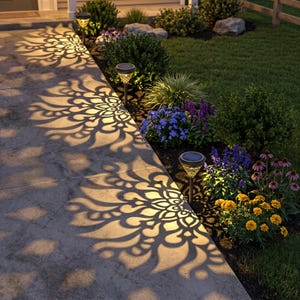 May include: Three solar-powered garden stake lights with dark metallic finishes and textured glass tops, positioned along a stone walkway, casting intricate, large, floral-shaped light patterns onto the concrete surface in a dark outdoor setting.
