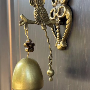 May include: A brass-toned metal wall-mounted door bell featuring a detailed owl perched on a branch. The owl is attached to a decorative backplate. A small flower charm connects the bell to the branch, and a wavy metal pull chain hangs below.