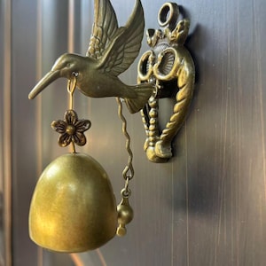 May include: A brass-toned metal door bell featuring a sculpted hummingbird in flight. The bird holds a small flower-shaped charm attached to a smooth, rounded bell. A decorative, ornate mounting bracket with a circular loop secures the piece.