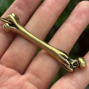 May include: A brass-colored bone-shaped pendant with a small loop at one end. The pendant has a detailed, realistic design, with visible ridges and curves. The overall aesthetic is antique or vintage.