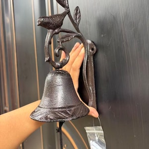 May include: A dark brown cast iron wall-mounted bell featuring a decorative bird perched on a branch with leaves. The bell has a flared rim with a textured pattern and hangs from a curved bracket. A black pull cord hangs from the center.