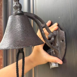 May include: A black cast iron wall-mounted bell featuring a textured, conical shape and a decorative curved bracket. A black rope pull hangs from the center of the bell, which is designed to be attached to a flat surface with visible screw holes.