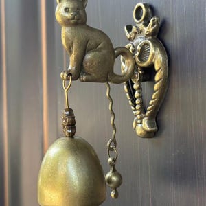 May include: A wall-mounted door bell featuring a bronze-toned cat figure sitting atop a decorative bracket. A small, smooth, dome-shaped bell hangs from the cat's front paw by a thin metal loop, while a wavy metal chain with a small sphere hangs below.