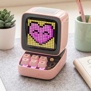 Puede incluir: Un altavoz rosa de estilo retro con una pantalla que muestra un corazón pixelado. El altavoz tiene una base tipo teclado con teclas iluminadas. Una pequeña suculenta en una maceta blanca y un bolígrafo en una taza verde están en el fondo.