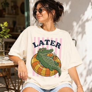 Könnte beinhalten: Cremefarbenes T-Shirt mit dem Wort "LATER" in Marineblau über einem Cartoon-Alligator, der eine Sonnenbrille trägt und auf einem gelb-orangenen Schwimmreifen schwimmt. Der Alligator hält ein Getränk. Hinter dem Text befinden sich rosa vertikale Streifen.