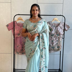 Peut inclure: Un sari bleu clair avec des broderies et des sequins détaillés. Le sari présente un motif floral et de plumes de paon sur la bordure. Deux chemisiers brodés sont suspendus sur un portant derrière le sari.