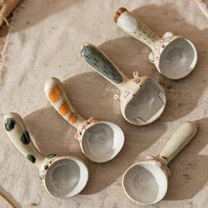 May include: A collection of five ceramic spoons with animal-themed designs. Each spoon features a different animal, with details like spots, stripes, and ears. The spoons are beige with colorful accents and a rounded bowl.