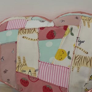 May include: A heart-shaped patchwork cushion featuring various fabric patterns including pink stripes, light blue polka dots, red strawberries, yellow lemons, and a tiger print with the word tiger written in black cursive script on a white background.