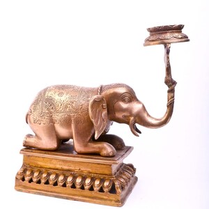 May include: A bronze elephant sculpture kneeling on a rectangular base, with an ornate design on its back and a curved trunk. A decorative stand extends from the elephant's back, topped with a small, round platform. The sculpture is a decorative item.