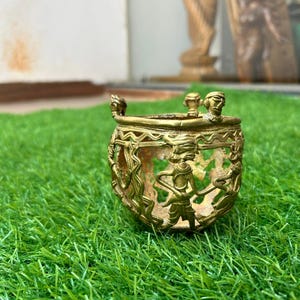 Peut inclure: Petit récipient en laiton antique avec des motifs découpés complexes représentant des personnages dansant ou en cérémonie. Le récipient a une forme arrondie avec un bord évasé et de petites figurines décoratives sur le dessus. Il est posé sur de l'herbe artificielle verte.