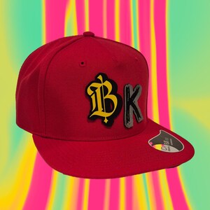 Könnte beinhalten: Rote Baseballkappe mit flachem Schirm. Auf der Kappe sind die Buchstaben "BK" in einem gelben und grauen Design auf der Vorderseite abgebildet. Der Hintergrund ist eine bunte Mischung aus Pink, Gelb und Grün.