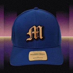 Könnte beinhalten: Blaue Baseballkappe mit einem goldfarbenen Buchstaben "M"-Aufnäher. Die Kappe hat einen gebogenen Schirm und ein Etikett mit der Aufschrift "Entworfen mit einem nachhaltigen Mindset mit recyceltem Polyester."