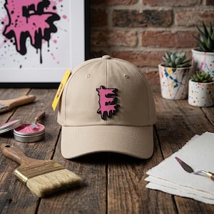 Könnte beinhalten: Eine beige Baseballkappe mit einem mittig platzierten, erhabenen Aufnäher des Buchstabens E im tropfenden rosa Farbdesign mit schwarzer Umrandung. Die Kappe verfügt über eine strukturierte Krone, einen gebogenen Schirm und ein kleines gelbes Etikett.