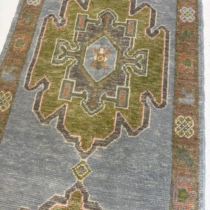 Könnte beinhalten: Ein rechteckiger Teppich mit einem hellblauen Feld und einem zentralen geometrischen Medaillon in Olivgrün und Anthrazit. Die Bordüre ist braun und mit kleinen Blumenmotiven in Hellblau, Grün und Orange verziert. Der Teppich hat einen Kurzflor.