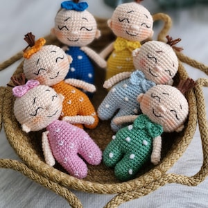 May include: A collection of seven handmade crochet dolls in a woven basket. Each doll has a smiling face, brown hair, and is wearing a different colored outfit with white polka dots. The outfits are pink, orange, green, blue, and yellow.