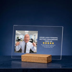 Op de afbeelding: Transparante acrylplaat met een houten voet. De plaat toont een foto van een man en de tekst "GOOD LUCK FINDING BETTER COWORKERS THAN US". De plaat is een afscheidscadeau.