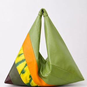 Puede incluir: Un bolso de mano de cuero con forma triangular y diseño de bloques de color. El bolso presenta paneles de color verde oliva, una franja naranja vibrante y una sección amarilla con un patrón tie-dye verde, con un detalle marrón oscuro.