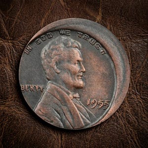 以下が含まれることがあります： 右を向いたエイブラハム・リンカーンの横顔が描かれた、1955年製の銅色のリンカーン・セント硬貨。硬貨の上端には「IN GOD WE TRUST」の文字、左側には「BERTY」、右側には「1955」という年号が刻まれています。