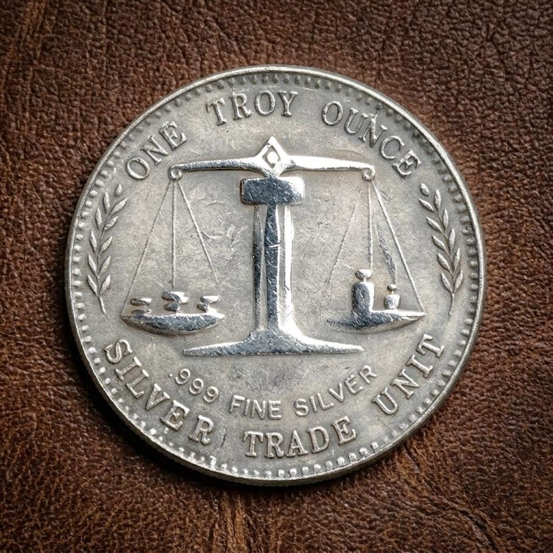 May include: A silver coin with the text "ONE TROY OUNCE" and "SILVER TRADE UNIT". The coin features a scale design with weights and a decorative border. The coin is .999 fine silver.